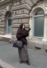 Giacca marrone oversize, guanti neri, gonna trasparente con motivo floreale, tacchi scuri e borsa, sullo sfondo un edificio con texture in pietra.