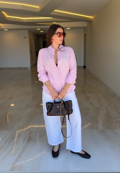 Mujer con una camisa rosa, pantalones blancos y zapatos negros, sosteniendo un bolso negro de mano, de pie en un pasillo moderno con suelos de mármol.
