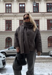 Ruda vilnonė striukė su aukštu apykakle ir dideliais mygtukais, derinama su pilkomis kelnėmis ir juoda rankine. Fone matoma sniego padengta gatvė.