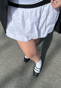 Gonna bianca plissettata con una cintura nera; abbinata a scarpe nere e calzini bianchi. Sullo sfondo si vede una superficie pavimentata.
