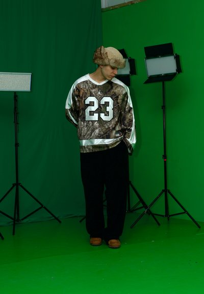 Camisa de gran tamaño con estampado de camuflaje y número 23, pantalones negros y un sombrero de piel marrón claro. Luces de estudio en trípode sobre un fondo verde.