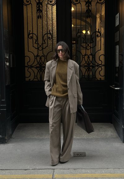 Traje beige claro con pantalones de pierna ancha, un suéter verde oscuro debajo y gafas de sol de gran tamaño. Sostiene un maletín marrón oscuro.