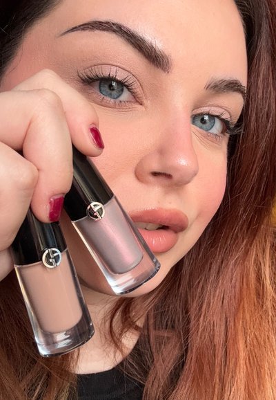 Dos botellas de esmalte de uñas de vidrio con tapones negros, una nude y otra rosa iridiscente, levantadas frente a una vista de primer plano del rostro de una persona.