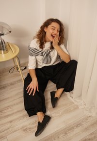 Haut blanc à rayures noires drapé au cou, pantalon ample noir et chaussures noires sans lacets, assise dans une pièce lumineuse avec un sol en bois clair.