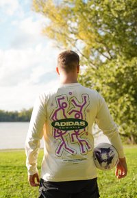 Μακρυμάνικο κρεμ πουκάμισο με μωβ γραφικό σχέδιο και λογότυπο "ADIDAS ORIGINALS" στο πίσω μέρος; μαύρη βερμούδα και μια μπάλα του ποδοσφαίρου στο χέρι.