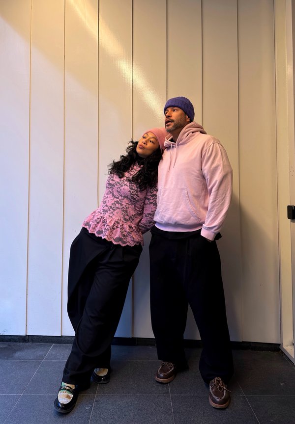 Sieviete rozā mežģīņu topā un cepurītē atbalstās pret vīrieti rozā džemperī ar kapuci un purpura cepurīti, abi tērpušies melnās biksēs un stāv pie baltas sienas.