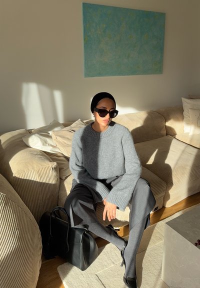 Suéter gris de punto con mangas extragrandes, pantalones negros y un bolso negro. La luz del sol proyecta sombras sobre un sofá beige texturizado y una alfombra clara.