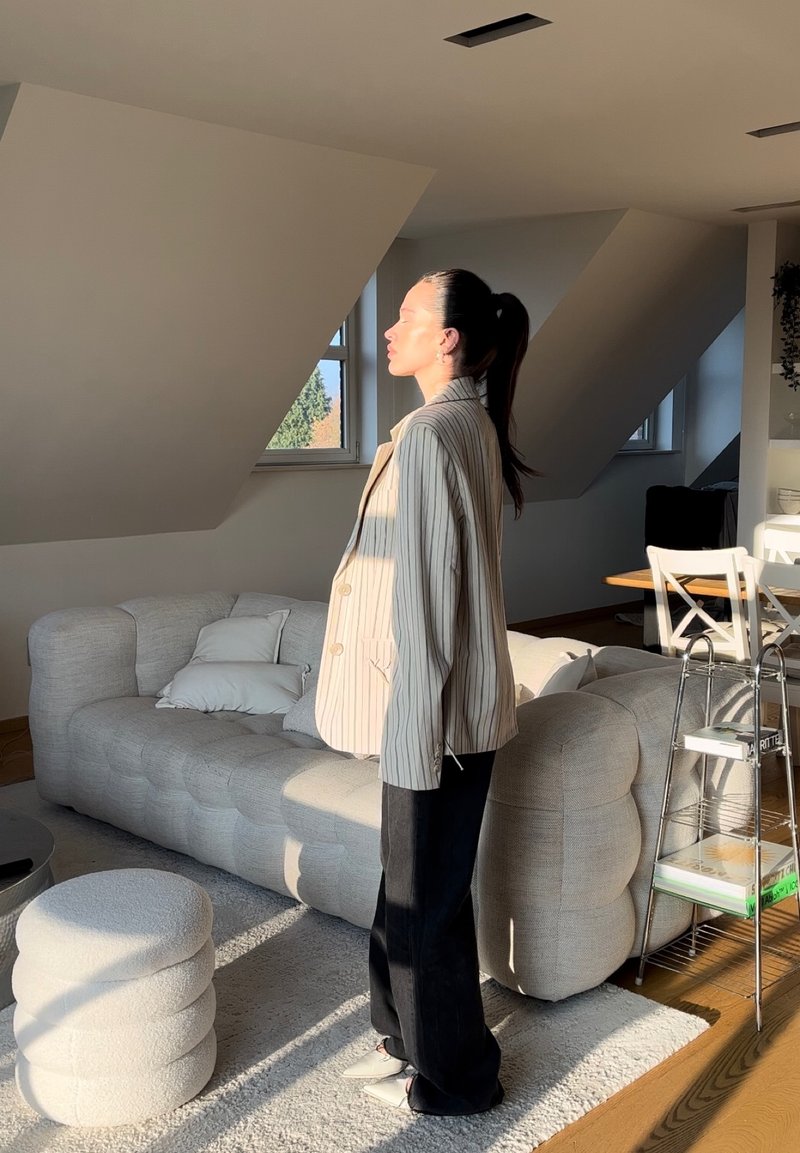 Ριγέ μπεζ σακάκι με δύο κουμπιά και φαρδιές πατιλέτες, συνδυασμένο με μαύρο παντελόνι και λευκά παπούτσια. Η πηγή φωτός δημιουργεί απαλές σκιές σε ένα μίνιμαλ εσωτερικό.