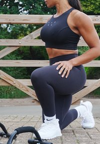 Černá sportovní podprsenka s logem, doplněná o černé legíny s vysokým pasem a bílé sportovní boty. Hladká látka, přiléhavý střih a čisté linie.