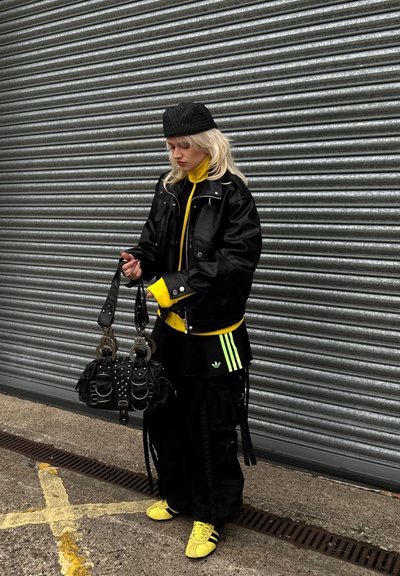 Chaqueta de cuero negra sobre un abrigo amarillo brillante con cremallera, falda negra con rayas verdes, bolso negro con tachuelas y zapatos amarillos.