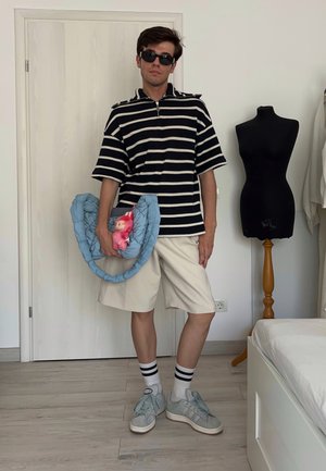 Camisa polo a rayas en blanco y negro, pantalones cortos beige, bolsa de tela azul con una muñeca rosa, zapatillas grises y gafas de sol negras sobre un fondo sencillo.