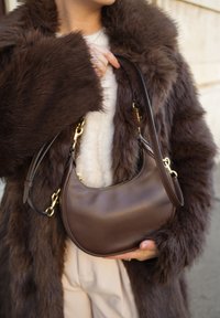 Bruine leren handtas met een afgerond ontwerp, voorzien van een ritssluiting en goudkleurige accenten. Getoond naast een bontjas.