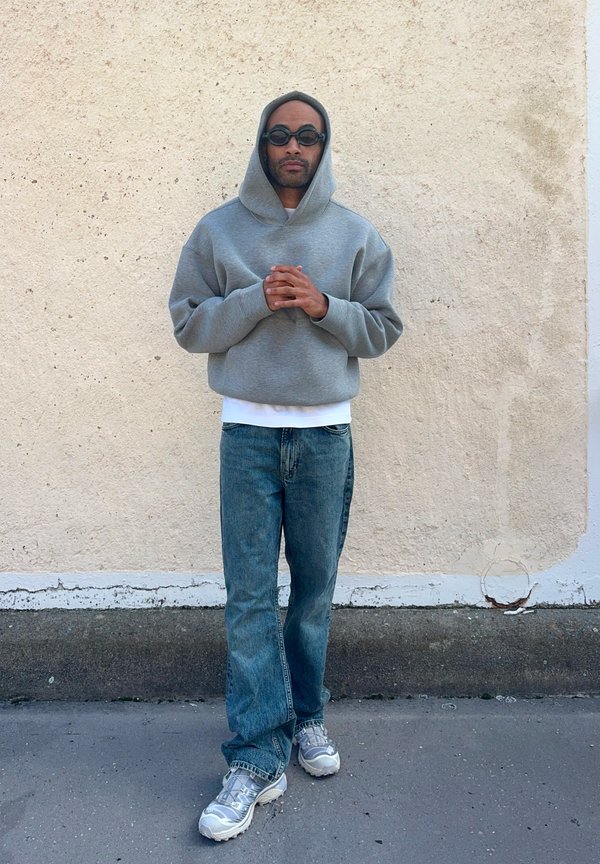 Grå hettegenser, hvit skjorte under, blå jeans og sølvfargede joggesko. Stoffet er mykt, med en avslappet passform og uformelt design.