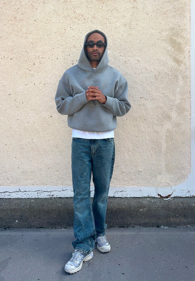 Grijze hoodie, witte shirt eronder, blauwe jeans en zilveren sneakers. De stof is zacht, met een relaxte pasvorm en een casual ontwerp.