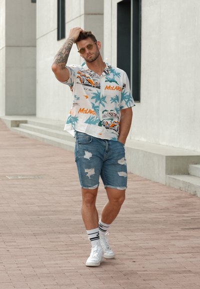 Camisa de manga corta blanca con estampado tropical en tonos azul y naranja, pantalones cortos de mezclilla desgastados, zapatillas blancas y calcetines a rayas.