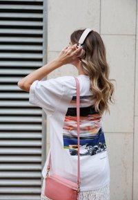 Λευκό μπλουζάκι με πολύχρωμο γραφικό σχέδιο ενός αγωνιστικού αυτοκινήτου. Συνδυάζεται με μια ανοιχτή ροζ τσάντα ώμου και ακουστικά. Ορατά κυματιστά μαλλιά.
