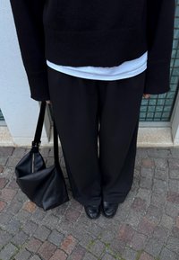 Άτομο που φοράει μαύρο πουλόβερ, λευκό πουκάμισο, μαύρο παντελόνι με φαρδιά μπατζάκια, μαύρα παπούτσια, κρατώντας μια μεγάλη μαύρη τσάντα, στέκεται σε πλακόστρωτο δρόμο.