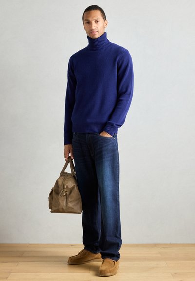 Suéter de cuello de tortuga navy, con puños y dobladillo acanalados; jeans de mezclilla oscura; bolso de cuero marrón; zapatos tipo slip-on en color beige; fondo liso con suelo de madera.