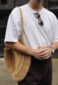 Rund, vävd stråväska i beige färg, placerad tillsammans med en vit t-shirt och mörka byxor, hålls av en person med en klocka.
