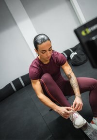 Athletic shoes with light grey and pink accents, deep burgundy fitted shirt and leggings, and a floral tattoo on the arm. Person tying shoelaces.