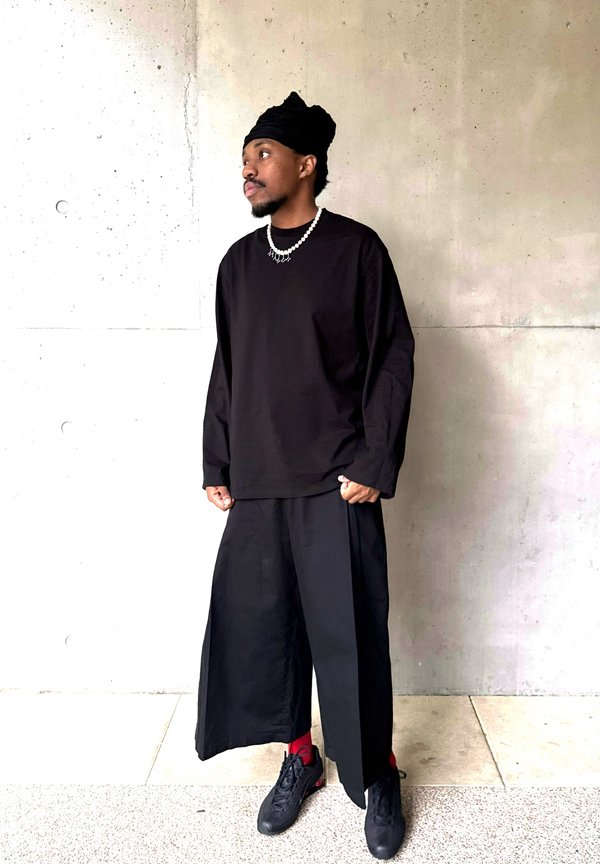 Homme portant une chemise noire à manches longues, un pantalon large noir, un foulard noir sur la tête, un collier de perles, des chaussettes rouges et des baskets noires, debout contre un mur gris.