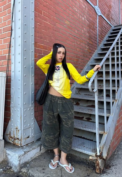 Joven mujer con camiseta amarilla de manga larga y pantalones de camuflaje posa junto a unas escaleras metálicas oxidadas y una pared de ladrillo rojo, llevando un bolso negro de hombro.