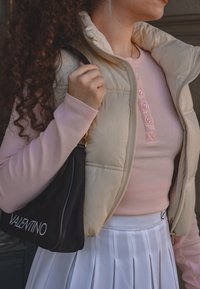 Kreminė pūkuota liemenė su šviesiai rožine raukšlėta ilgų rankoviu palaidine su mygtukais, derinama su klostuota balta sijonu ir juoda Valentino rankine.
