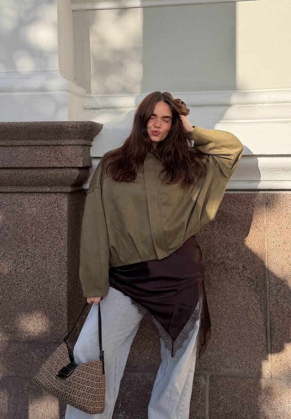 Joven con cabello largo y oscuro, con una chaqueta oliva de gran tamaño y pantalones blancos, sosteniendo un bolso estampado, de pie frente a una pared de piedra y blanca.