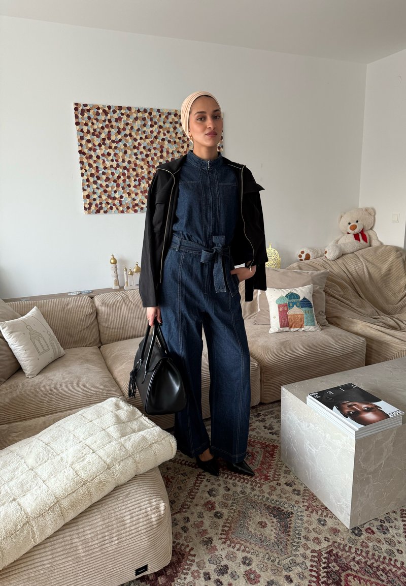 Femme portant une combinaison en denim et une veste noire, tenant un sac à main noir, debout dans un salon avec des meubles beiges et un tapis à motifs.