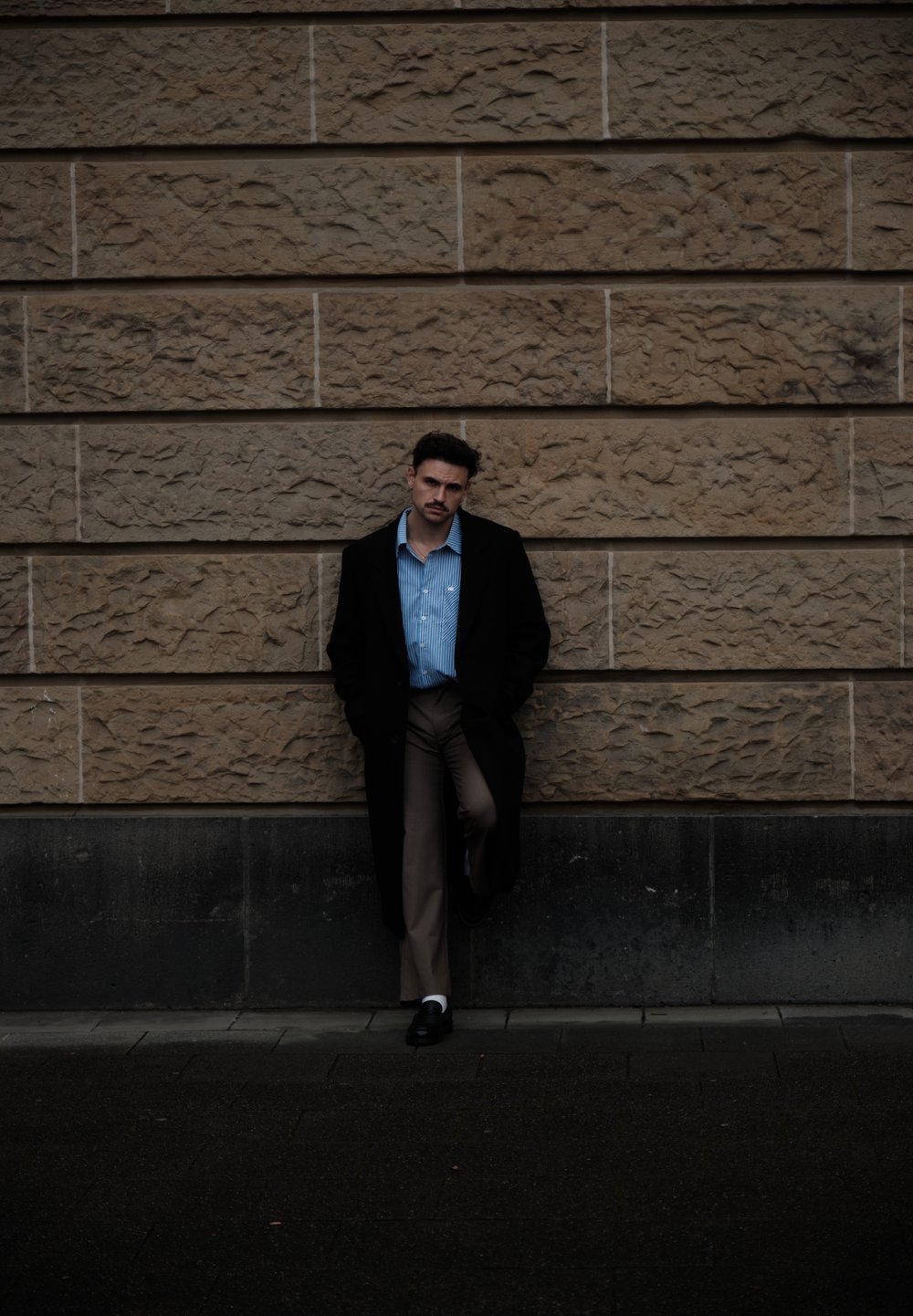 Czarny płaszcz, jasnoniebieska koszula w paski i beżowe spodnie. Stoi na tle teksturowanej kamiennej ściany. Ciemna podłoga, subtelne kontrasty świetlne.