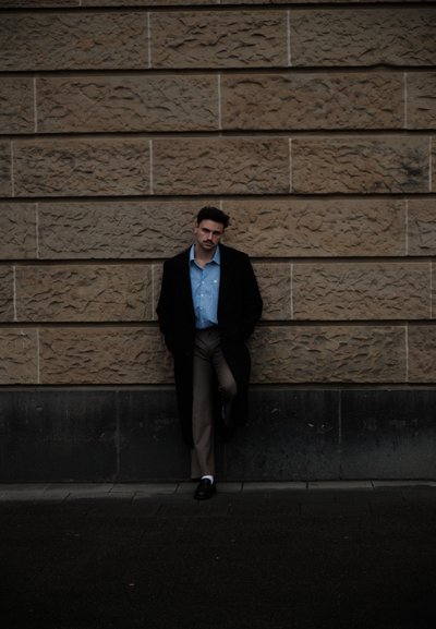 Abrigo negro, camisa azul claro a rayas y pantalones beige. De pie frente a una pared de piedra texturizada. Suelo oscuro, contrastes sutiles de iluminación.