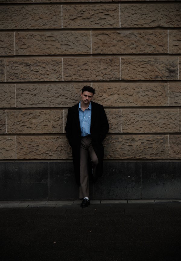 Schwarzer Mantel, hellblau gestreiftes Hemd und beige Hose. Stehend vor einer strukturierten Steinmauer. Dunkler Boden, dezente Lichtkontraste.