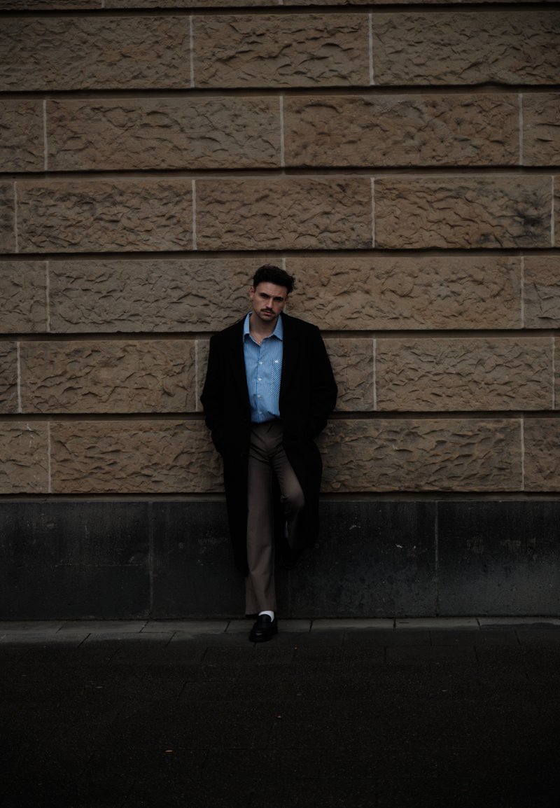 Crni kaput, svijetloplava košulja s prugama i bež hlače. Stojeći naslonjen na teksturirani kameni zid. Tamna podloga, suptilni kontrasti osvjetljenja.