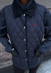 Veste matelassée bleu marine avec col en velours côtelé, boutons-pression et deux poches avant, avec un petit logo rouge sur le côté gauche.