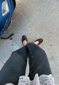 Tamsiai mėlynos džinsai, rudos plokščios bateliai su dekoratyvine kaspinu ir balta marškinėlių su nėriniais viršūnė matomi ant pilko akmens grindinio.