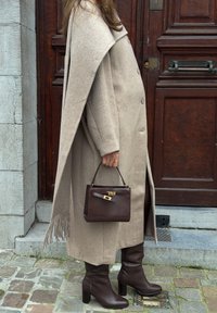 Άτομο που φοράει μακρύ παλτό σε μπεζ χρώμα, σκούρα καφέ μπότες με τακούνι, κρατώντας μια τσάντα χειρός σε σκούρο καφέ χρώμα, στέκεται μπροστά από μια ξύλινη πόρτα σε πλακόστρωτο.