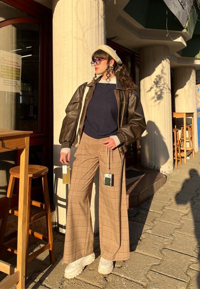 Joven mujer con pantalones marrón a cuadros de pierna ancha, suéter negro, chaqueta de cuero marrón oscuro, boina beige y zapatillas blancas, de pie fuera de un café sosteniendo una botella.
