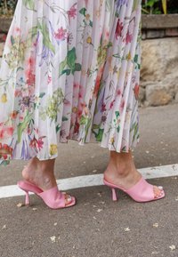 Mule rosa con punta aperta, dalla finitura liscia e opaca e un tacco corto e sottile, abbinati a una gonna a pieghe con motivo floreale.