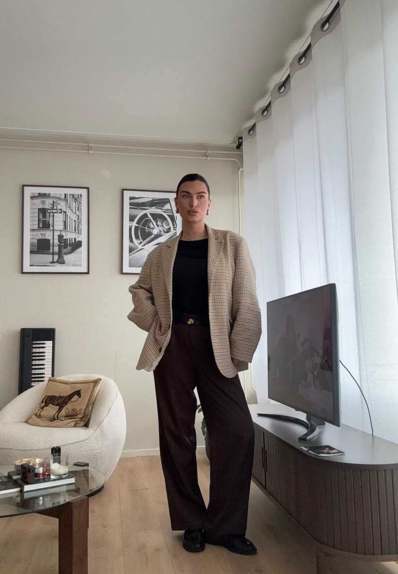 Femme en blazer beige à carreaux et pantalon foncé debout dans un salon moderne avec un fauteuil blanc, une table en verre, une télévision et des photos en noir et blanc encadrées.