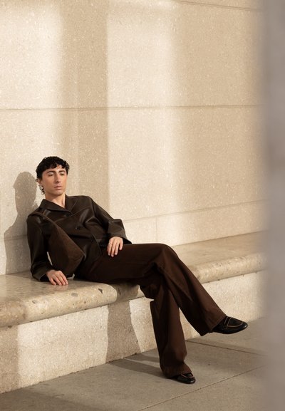 Persona joven con cabello rizado, vistiendo ropa y zapatos de color marrón oscuro, recostada en un banco de piedra contra una pared texturizada beige a la luz del sol.