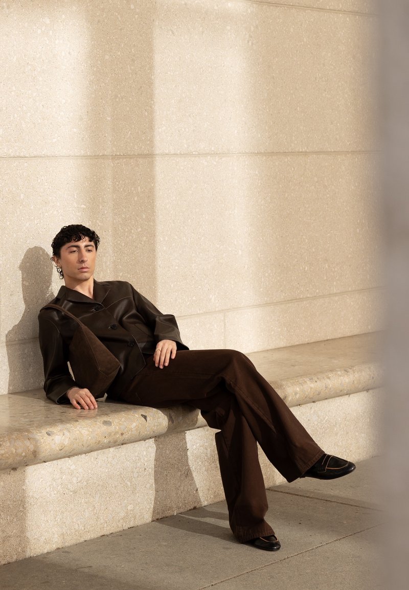 Jeune personne aux cheveux bouclés portant une tenue marron foncé et des chaussures, appuyée en arrière sur un banc en pierre contre un mur beige texturé au soleil.