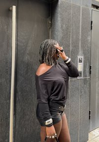 Personne aux cheveux argentés tressés, portant un haut noir décolleté et un short en cuir, ajustant ses lunettes de soleil tout en se tenant près d'un mur de pierre sombre et d'une porte.