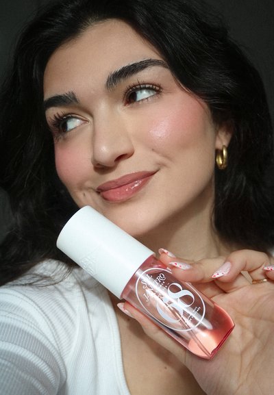 Joven con cabello oscuro y pendientes de aro dorados sostiene cerca de su rostro una botella rosa de cuidado de la piel etiquetada como "SALT JANEIRO 08", sonriendo.