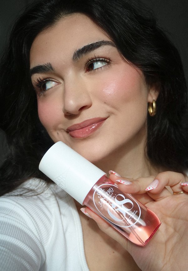 Joven con cabello oscuro y pendientes de aro dorados sostiene cerca de su rostro una botella rosa de cuidado de la piel etiquetada como "SALT JANEIRO 08", sonriendo.