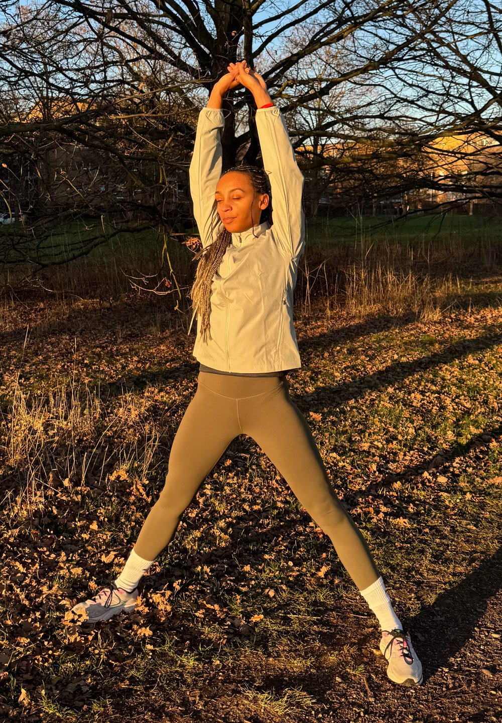 Vêtements de sport : veste zippée gris clair, couche de base noire, leggings olive. Décor extérieur avec des feuilles tombées, personne s'étirant sous le soleil.