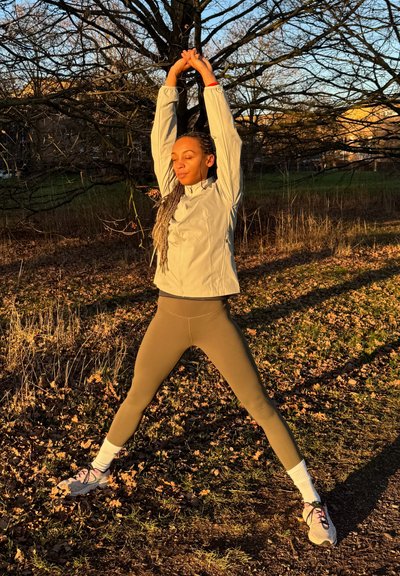 Ropa deportiva: chaqueta con cremallera gris claro, capa base negra, leggings color oliva. Entorno al aire libre con hojas caídas, persona estirándose bajo la luz del sol.