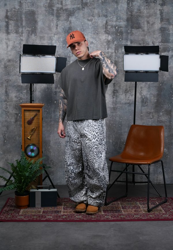 Joven con los brazos tatuados, que lleva una gorra marrón, una camisa gris extragrande y pantalones con estampado de leopardo, está de pie entre dos luces en un estudio.