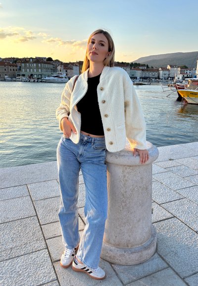 Chaqueta de mezcla de lana crema con botones dorados, top negro corto, jeans azul claro y zapatillas blancas con acentos oscuros, de pie junto a un pilar de piedra.
