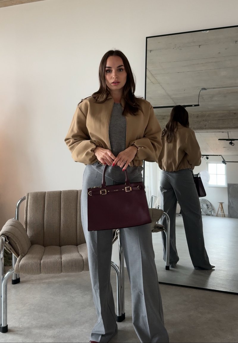 Femme en veste beige et pantalon gris tenant un sac à main rouge foncé, debout devant une chaise beige rembourrée et un grand miroir mural.