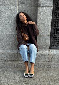 Kobieta z kręconymi włosami siedzi na kamiennym brzegu przy szarym murze, ubrana w brązową marynarkę, jasnoniebieskie jeansy, wzorzyste baleriny oraz brązową torebkę na ramię.